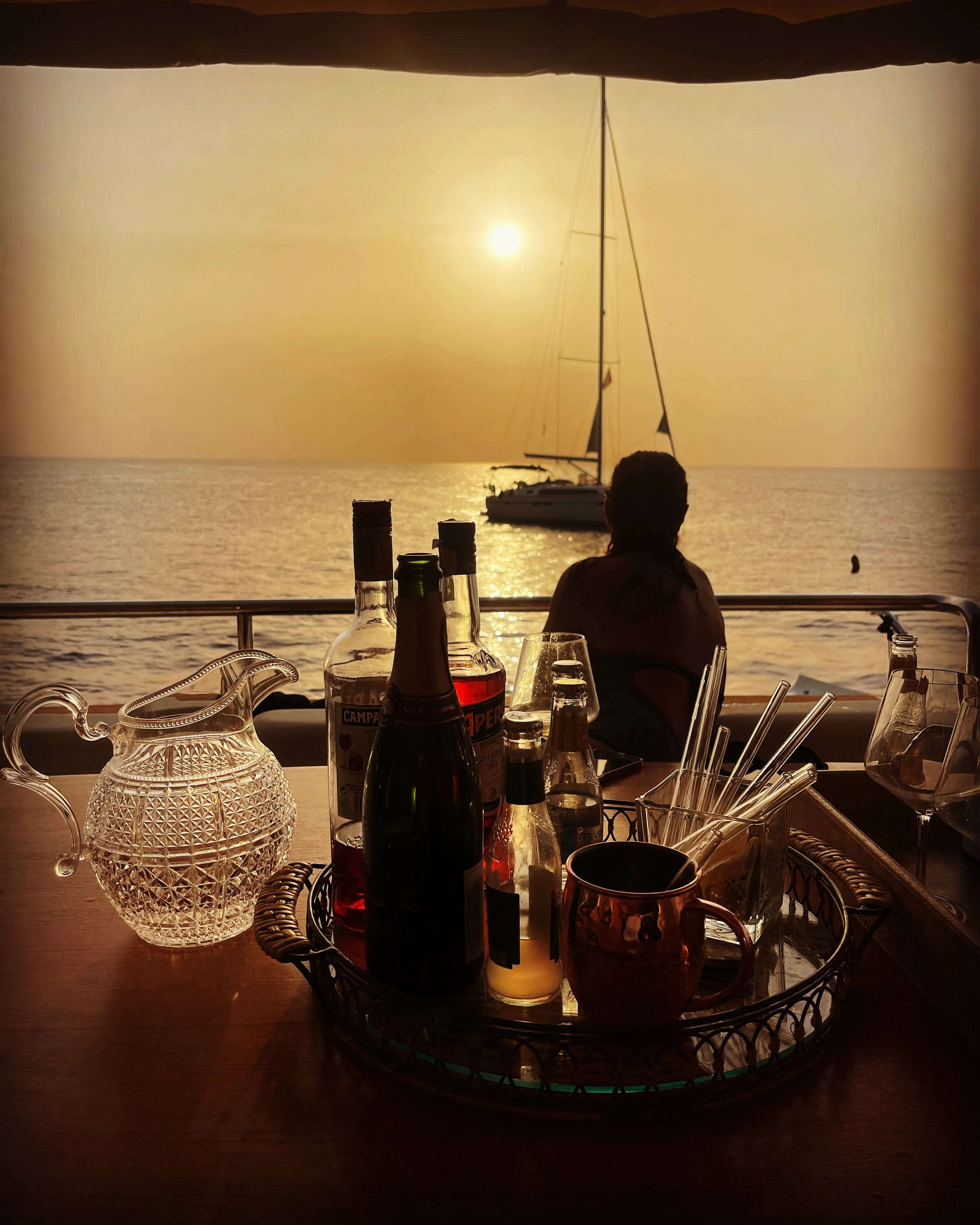 The drinks tray on the aft deck table, a sailing boat backlit at sunset