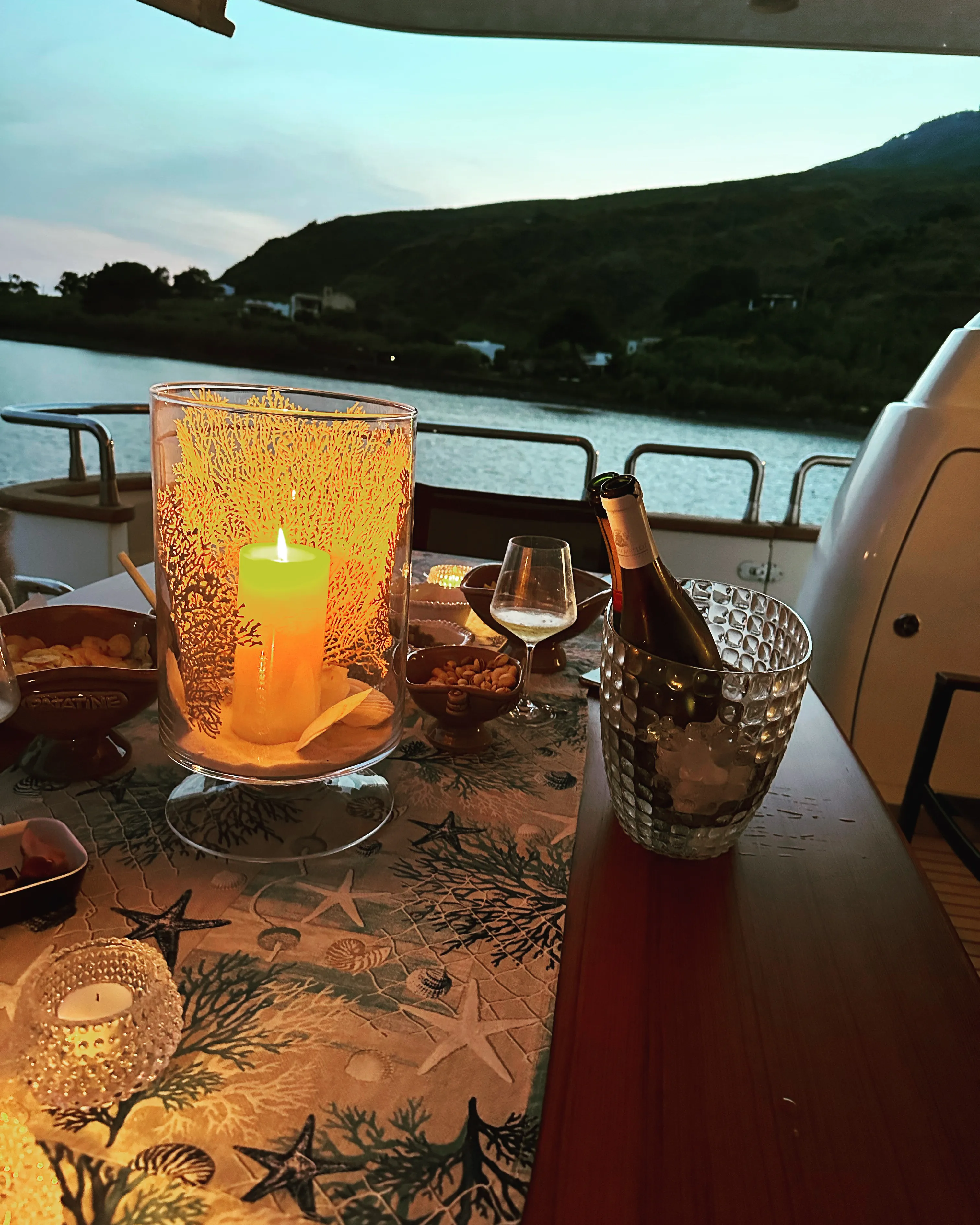 A lit candle on the aft deck table and a bottle on ice, the coast at dusk